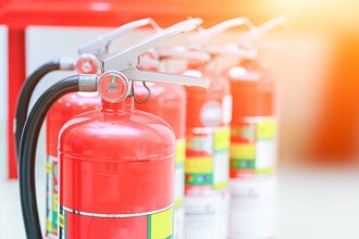 Mehrere Feuerloescher vor einem weiß-rot-orangen Hintergrund. | © A Stockphoto - AdobeStock