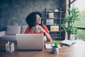 Porträt einer verträumten Frau, die am Schreibtisch in einem modernen Büro sitzt mit einem heißen Getränk in der Hand aus dem Fenster schaut | © deagreez - AdobeStock